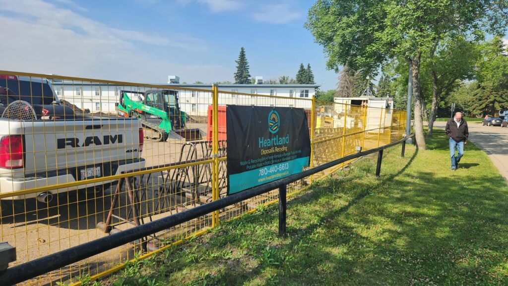 A fenced construction site with a RAM truck, mini excavator, and a Heartland Fence Rentals banner. A man walks on a sidewalk beside green grass and trees under a partly cloudy sky.