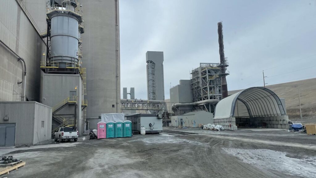 An industrial facility with large concrete structures and silos, portable toilets, a white canopy shelter, a few vehicles, and scattered construction materials on a muddy, overcast day.