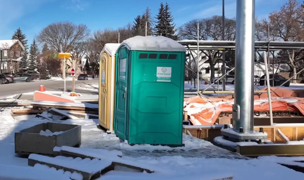 A green portable toilet stands on a snowy construction site, surrounded by scattered materials and snow. Leafless trees and residential houses are visible in the background under a clear blue sky.