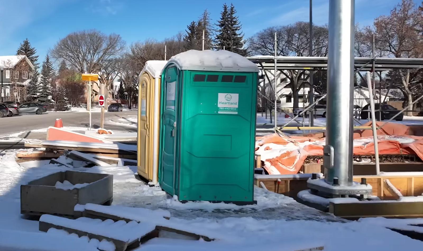 A green portable toilet stands on a snowy construction site, surrounded by scattered materials and snow. Leafless trees and residential houses are visible in the background under a clear blue sky.