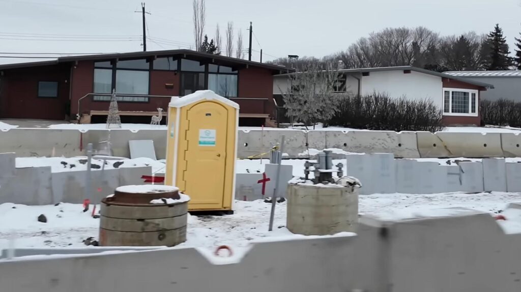 A snowy construction site with a yellow portable toilet in front of residential houses. Concrete barriers and construction materials are scattered across the site. Bare trees and overcast sky in the background.