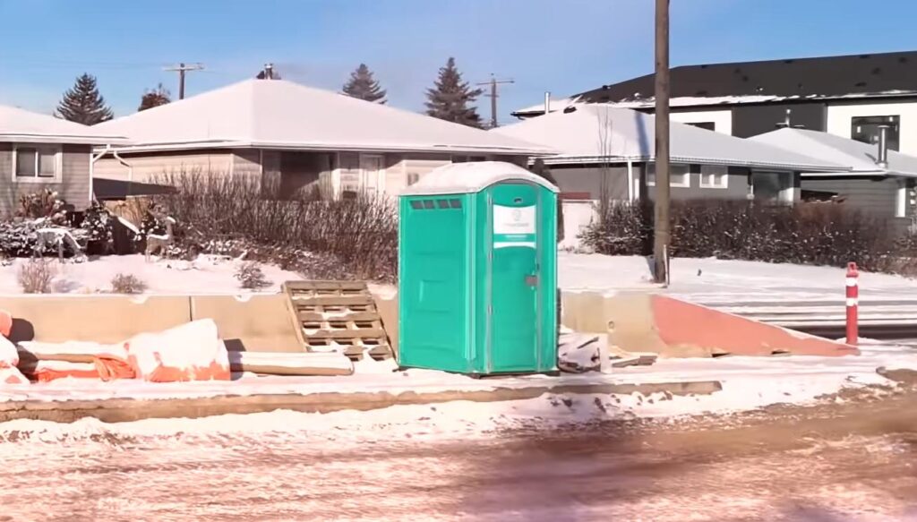 A green portable toilet stands on a snowy roadside in front of houses, with construction materials and barriers scattered nearby on a cold, sunny day.