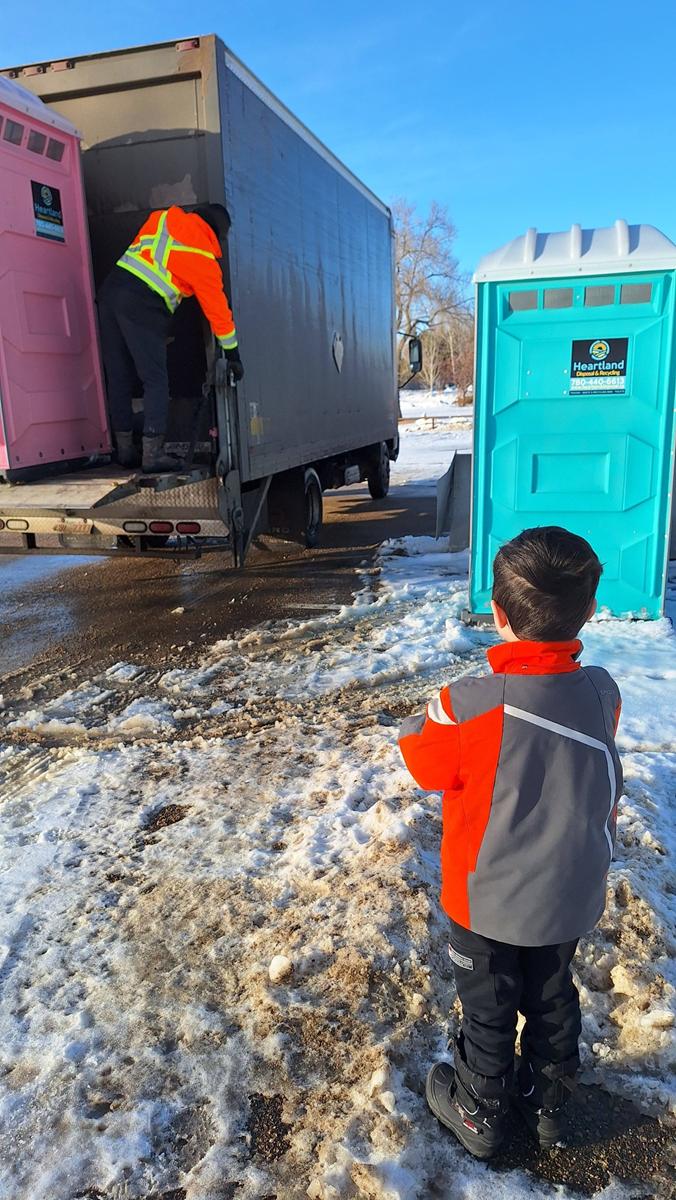 A child in a red and gray winter coat stands on snowy ground, watching a worker wearing a safety vest unload a pink portable toilet from a truck near a blue portable toilet.