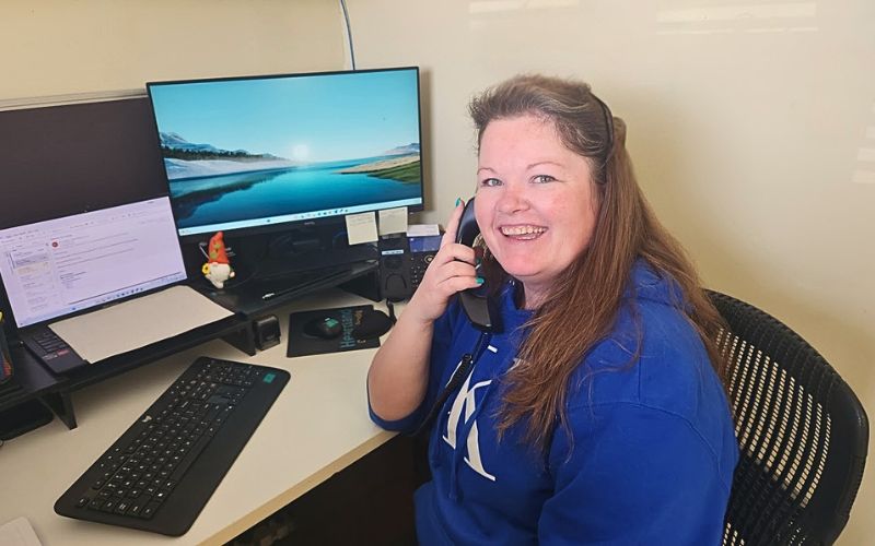 A smiling woman in a blue hoodie sits at a desk with computer monitors, holding a phone to her ear, appearing to work in an office environment.