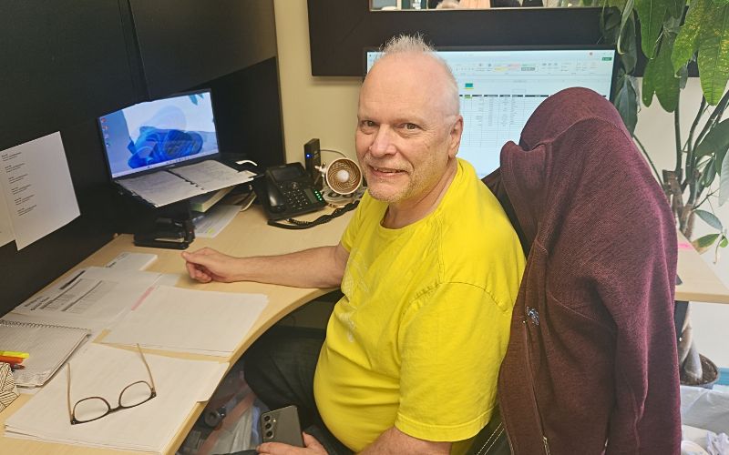 A man in a yellow shirt sits at his desk, smiling at the camera. Papers, a phone, glasses, and a laptop with a blue image are on the desk. A maroon jacket is draped over his chair. A plant is visible nearby.