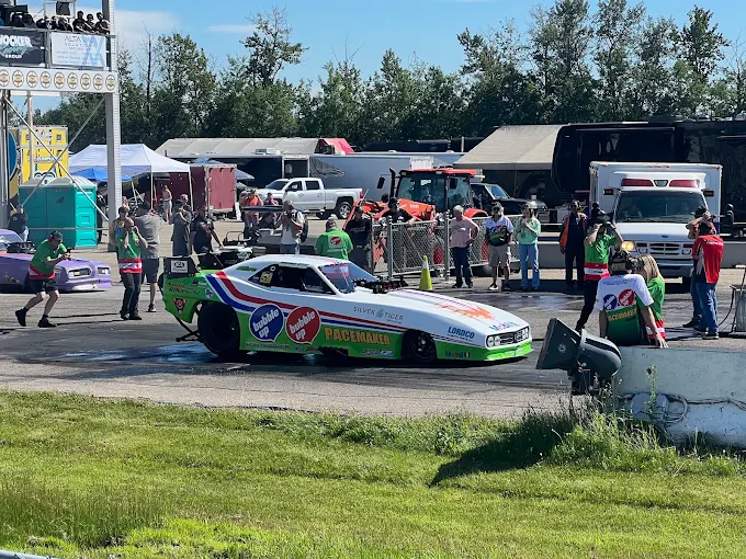 A colorful drag race car waits at the starting line as people gather around, some in safety gear. Spectators and vehicles are visible in the background under a sunny sky.