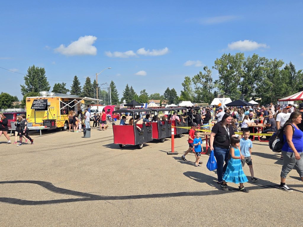 A crowd enjoys a sunny outdoor food truck festival with families walking, people eating at picnic tables, and colorful trucks lined up in the background.