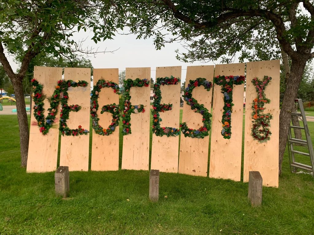 Large wooden boards display the word VEGFEST spelled out with colorful flowers and greenery, set outdoors between two trees.